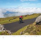 März. Ein Radfahrer auf einer Bergstraße, umgeben von grünen Wiesen und Wolken, klarer Himmel. Logo: Qualdich.