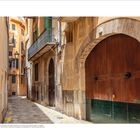 Text "Das Zentrum", schmale, enge Gasse in mediterraner Altstadt, helle Gebäude mit grünen Fensterläden und Holztüren.