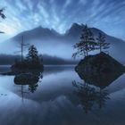 Keine Texte vorhanden. Eine ruhige Landschaft mit einem See, Inseln mit Bäumen, Bergen und Nebel, in blauem Licht getaucht.