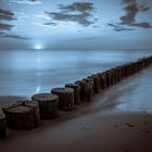 Holzpfeiler führen ins Meer unter einem blau getönten Himmel mit Wolken, weit weg ein heller Horizont.