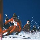 Ein Skifahrer in orangener Kleidung fährt durch den Schnee. Sein Helm hat ein gemustertes Design. Zuschauer im Hintergrund.