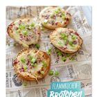 "Flammkuchen-Brötchen, 2 Portionen." Vier Brötchenhälften mit Käse, Speck und Frühlingszwiebeln auf bedrucktem Papier.