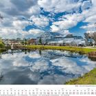 März 2027 Kalender mit einem modernen Glasgebäude am See. Besucher spazieren, blauer Himmel mit Wolken spiegelt sich im Wasser.