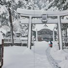Schneebedeckte japanische Torii und ein Mensch räumt Schnee, Bäume und ein Gebäude im Hintergrund.