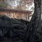 Ein modernes Haus mit großen Fenstern auf einem Felsen, umgeben von Bäumen mit herbstlichem Laub. Rauch steigt auf.