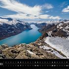 Norwegen, 03. Besseggen, Jotunheimen, Vågå. Ein blauer See, umgeben von schneebedeckten Bergen unter klarem Himmel. Kalender unten.