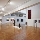 Keine lesbaren Texte vorhanden. Eine Kunstgalerie mit Skulpturen, Gemälden an weißen Wänden und einem Holzboden.