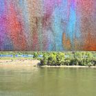 "OEVG KAGU" in Blau-Gelb auf Containern, bunte abstrakte Illustration oben, Fluss und Ufer mit Bäumen unten.