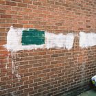 Eine Ziegelwand mit weiß übermalten Flächen und einem grünen Rechteck. Auf dem Boden liegt ein Papiersack von Lidl.