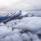 Eine verschneite Bergspitze erhebt sich majestätisch über dichten Wolken im Hintergrund; die Atmosphäre wirkt ruhig.