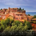 "Rote Ockerhäuser von Roussillon 4." Rote Häuser auf einem Hügel, umgeben von grünen Bäumen unter blauem Himmel.