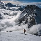 Ein Bergsteiger in roter Jacke erklimmt eine schneebedeckte Bergflanke, umgeben von Wolken und hohen Gipfeln in der Ferne.