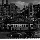Text: "Münchener Kindl." Eine belebte Szene in einer Stadt mit Straßenbahnen, Fußgängern und Autos, umgeben von prächtigen Gebäuden.