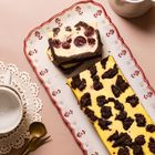 Ein rechteckiger Kuchen mit dunklen Schokostücken auf hellem Belag, daneben Tasse, Löffel und Gabel auf Häkeldeckchen.