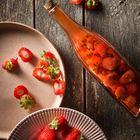 Erdbeeren auf zwei Tellern, eine Glasflasche mit roten Flüssigkeit und in Scheiben geschnittenen Erdbeeren auf Holztisch.