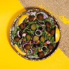 Ein Teller mit Falafelbällchen, Rucola, Zwiebeln, Zitronenscheiben und einem gelben Hintergrund, teils mit Linsen bedeckt.