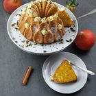 Ein Gugelhupf-Kuchen mit Sahnetupfen auf einer weißen Platte, daneben Kürbisse und eine Zimtstange. Ein Stück Kuchen liegt auf einem Teller.