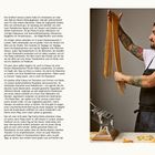 Einleitung: Ein tätowierter Mann in Schürze hält lange, frische Nudeln. Neben ihm eine Nudelmaschine und Pasta auf dem Tisch.
