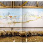 Ein Gemälde an einer Wand zeigt eine einfache Landschaftsskizze. Davor stehen Schafe auf Stroh in einem Stall.