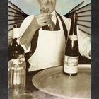 Der Text lautet "GASTFREUNDSCHAFT". Ein Kellner in Schürze hält ein Glas Wein, Flasche und Gläser sind auf einem Tablett.