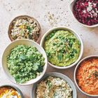 Verschiedene bunte Schüsseln mit Dips: grün mit Erbsen, rot mit Roter Bete, orange mit Möhren, und weitere mit frischen Kräutern.