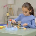 Mädchen in blauem Pullover spielt mit einer Spielzeugküche, umgeben von bunten Bechern auf einem Tisch in einem gemütlichen Raum.