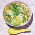 Ein Teller mit Nudelsuppe, grünem Gemüse und Tofu. Daneben liegt ein gelber Löffel auf einer strukturierten Oberfläche.