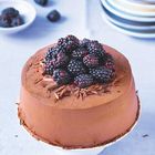 Ein großer, mit Schokolade überzogener runder Kuchen, garniert mit frischen Brombeeren und Schokoraspeln auf einem weißen Teller.