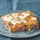 Ein Stück Kuchen auf einem Teller, bedeckt mit Mandelblättchen und Puderzucker. Der Hintergrund ist ein blauer Stoff.