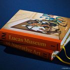"Lukas Museum of Narrative Art" auf zwei orangefarbenen Büchern. Oben ist ein Kunstwerk mit bunten Figuren.