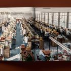Text "1970s" oben links. Große Werkhalle mit vielen Personen, die an langen Tischen arbeiten, wahrscheinlich Fabrikarbeit.