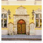 Nummer 26, Kalenderdaten für August 2027. Verzierter grüner Holztor in gelber Fassade mit steinernen Verzierungen.