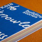 Text: "building stories". Ein blaues Buch mit farbig geschichteten Seiten auf einem orangen Hintergrund.