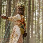 Keine Texte vorhanden. Eine Frau in einem dekorativen, floralen Kleid steht im Wald. Eine Leiter ist im Hintergrund.