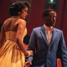 Keine sichtbaren Texte. Eine Frau im gelben Kleid hält den Arm eines Mannes im blauen Anzug in einem Theater.