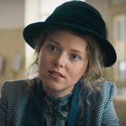 Eine Frau mit einem schwarzen Hut, lockigem Haar und einem karierten Outfit schaut aufmerksam. Der Hintergrund ist unscharf.