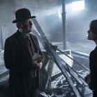 Ein Mann mit Melone hält einen Brief in einer düsteren, verfallenen Umgebung, spricht zu einer Frau.