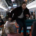 Ein Mann springt in einem modernen Zugabteil, während andere Passagiere überrascht schauen.