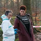 Zwei Personen im Gespräch im Wald. Eine im weißen Schutzanzug, die andere in einem dunklen Mantel. Absperrband im Hintergrund.