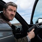 Ein Mann sitzt im Auto, hält eine Pistole und schaut ernst. Die Autotür ist offen, der Himmel ist bewölkt.