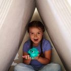 Ein Kind mit Zöpfen sitzt lächelnd in einer Zeltstruktur und hält ein leuchtendes blaues Spielzeug mit einem freundlichen Gesicht.