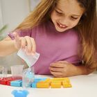Ein Mädchen in lila T-Shirt gießt blaue Flüssigkeit in eine Form. Bunte Formen und Verpackungen auf dem Tisch.