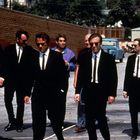 Mehrere Männer in schwarzen Anzügen und Sonnenbrillen gehen auf einer asphaltierten Straße. Eine rote Ziegelmauer im Hintergrund.