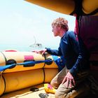 Ein Mann blickt von einem Schlauchboot aus aufs Meer, wo ein sinkendes Boot zu sehen ist.