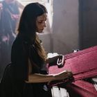 Eine Frau arbeitet konzentriert an einem traditionellen Webstuhl, mit rotem Stoff, in einem dimm beleuchteten Raum.