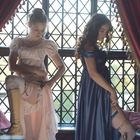 Zwei Frauen in eleganten Kleidern stehen an einem Fenster und schnallen sich Messer um die Oberschenkel.