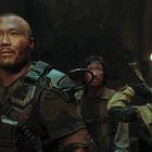 Text: "B01" an einer Wand. Drei Menschen in militärischer Ausrüstung sehen entschlossen aus.