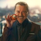 Ein Mann im Anzug hält eine kleine rote Daruma-Puppe in der Hand und lächelt. Im Hintergrund eine unscharfe Stadtansicht.