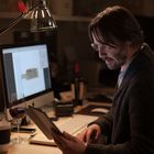 Ein Mann sitzt an einem Schreibtisch, liest auf einem Tablet. Ein Computerbildschirm und ein Glas Rotwein sind zu sehen.