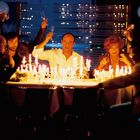 Mehrere Menschen heben Sektgläser um einen Tisch voller Kerzen. Die Szenerie wirkt festlich und lebhaft.